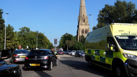 SALFORD, GREATER MANCHESTER, ENGLAND, UNITED KINGDOM - CIRCA SEPTEMBER, 2021: Ambulance van with blue emergency lights rushing by the church and through traffic lights at crossroads on Eccles Old Road