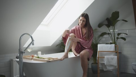 Young Beautiful Woman Shaving Leg In Pink Bathrobe In Modern Bathroom In House Care For The Skin And Preparing To Date Or Wedding She Using Shaving Cream And Razor Depilation Or Waxing Concept
