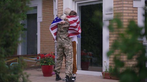 Wide shot joyful teenage boy dancing with American flag in slow motion standing on porch as happy military woman in camouflage uniform running to son hugging teenager. Excited family reunion outdoors