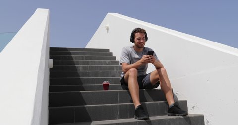 Podcast. Man listening to music, audiobook or podcast mobile phone app with headphones sitting on stairs. Healthy lifestyle person using smartphone on running break with morning red beet juice