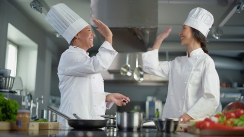 Restaurant Kitchen: Asian and Black Female Chefs Preparing Dish, Tasting Food and Doing High-Five in Successful Celebration. Two Professionals Cooking Delicious, Authentic Food, Healthy Meals Recipe