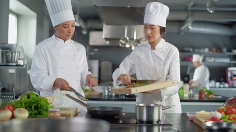Restaurant Kitchen: Portrait of Asian and Black Female Chefs Preparing Dish, Cutting Vegetables, Stirring Ingredients on the Frying Pan. Two Professionals Cook Delicious, Authentic Food, Healthy Meals