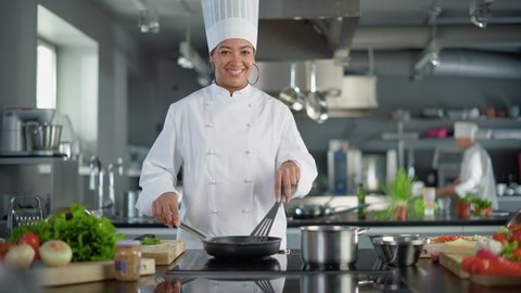 Restaurant Kitchen: Portrait of Black Female Chef in Action, Uses Pan to Cook Delicious, Traditional Authentic Food, Looks at Camera and Smiles. Healthy gourmet Dishes. Medium Wide Shot