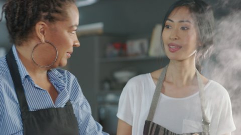 Restaurant Kitchen: Portrait of Black and Asian Female Chefs Talking, Preparing Dish, Tasting Food, Enjoying it. Two Professionals Cooking Delicious, Authentic, traditional Food, Healthy Meal Recipe