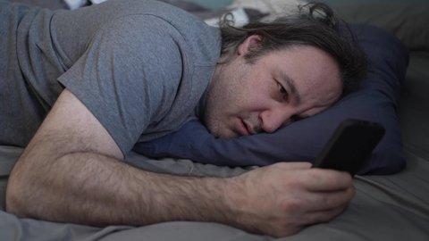 Man in his 40's using his cell phone in the bed and falling asleep all of a sudden. He is looking tired.