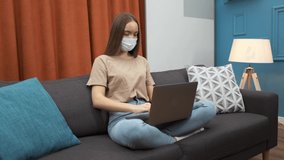 Sick young woman with protective mask coughing while working from home and using laptop at sofa. Video conference with webcam