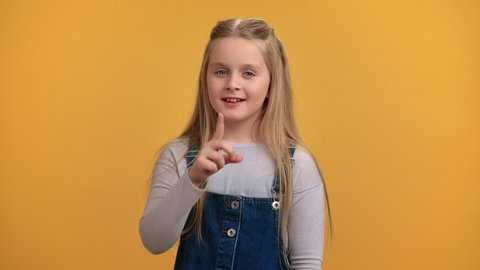 Portrait cute blonde baby girl posing with index finger on lips gesturing silence shhh noise mute isolated on orange. Cheerful female little kid playing performing quiet calm mysterious gesture