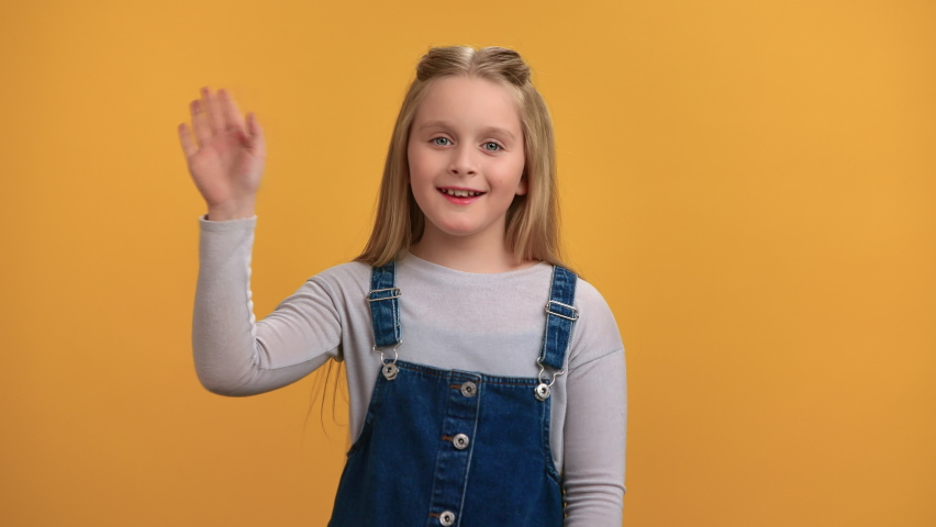 Portrait friendly smiling female kid waving hand with positive emotion posing isolated on orange studio. Happy cute little girl in denim sundress greeting welcome gesture congratulations her friends 