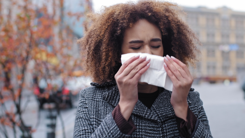 Sick ill african american woman with curly hair blowing runny nose into tissue paper outdoors cold allergies respiratory virus girl suffers from contagious rhinitis symptom autumn in city flu pandemic Royalty-Free Stock Footage #1086115232
