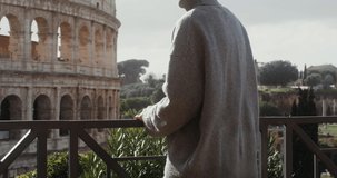 A young woman of European appearance in glasses, dressed in a sweater admires the Colosseum, standing on the observation deck on a sunny clear day. Italy Rome. Video in 4k, red komodo.