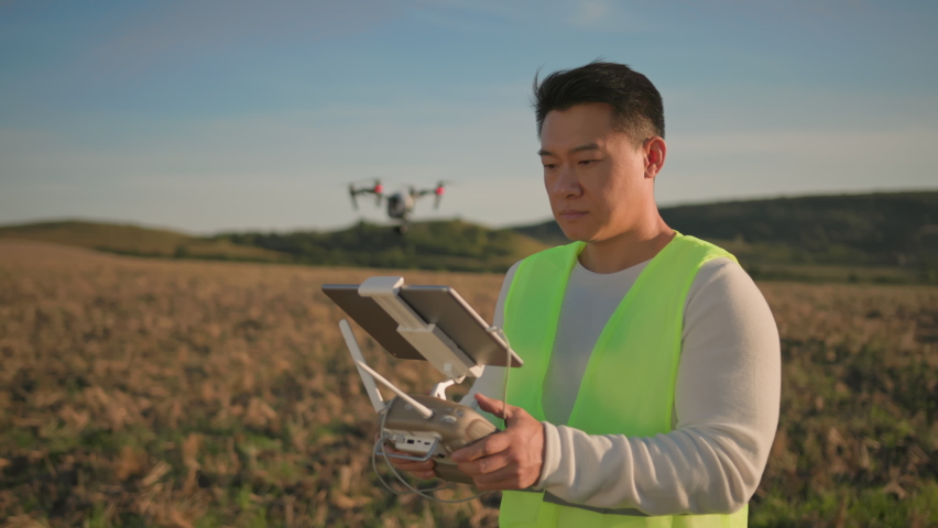 Industrial chinese worker wearing green vest, holding computer tablet drone controller and flying drone on the field. Agribusiness innovation. Technologies. Royalty-Free Stock Footage #1086116342