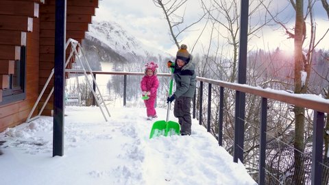 Snow shoveling from the terrace above the mountain lake. Children play with snow after a heavy snowfall. Smiling kids shoveling the snow from the terrace in sunny winter day.  