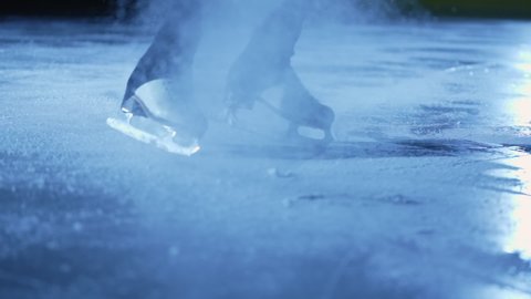 Detailed shot of women's legs in figure skating skates sliding on ice in an arena in dark with blue light. Woman slides on ice, splashing particles of sparkling ice into camera. Close up. Slow motion.