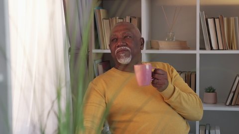 Enjoying life. Senior african man. Coffee break. Satisfied aged guy drinking hot beverage feeling good looking daylight window room interior.