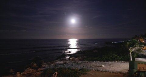 Beautiful view of bright moon in slightly cloudy sky above calm ocean and beach. Peaceful view of moonlight reflected on wavy sea near tropical coast. Night landscapes and holidays