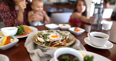 Female hand picking tortilla chip with cheese in slow motion from plate of chilaquiles with fried egg on top. Blurry kids and woman enjoying delicious traditional meal. Authentic Mexican cuisine