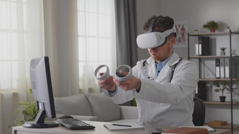 Young male doctor in white gown and VR headset sitting at desk in cabinet and use controller for virtual operation
