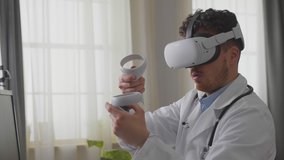 Young adult mane doctor in white gown and VR headset sitting at desk in cabinet and use controller for virtual operation