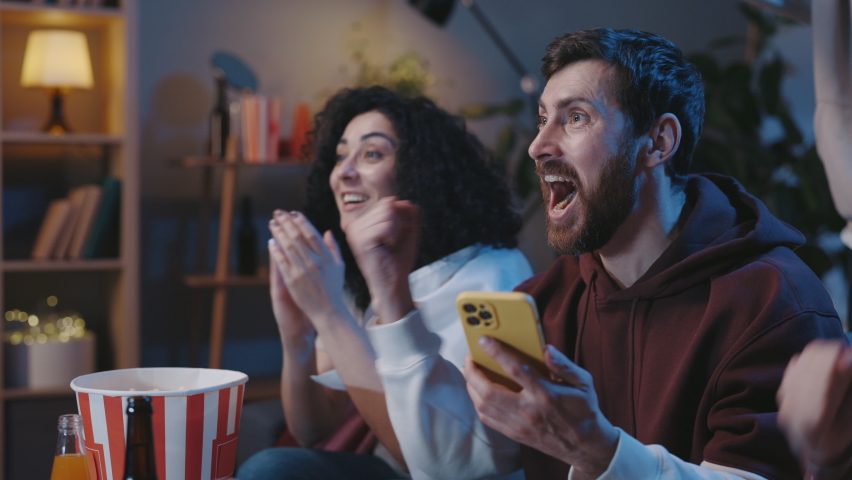 Enjoy favorite football club play. Soccer fans sitting on a couch and watching sports game on TV, while checking results at the smartphone at special app to online bet. Team wins championship concept | Shutterstock HD Video #1086139427