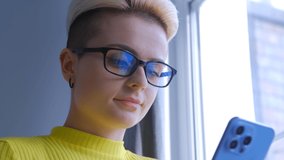 Young white woman typing message on phone. Stylish female with dyed short hair using modern smartphone for online communication. Tom boy person in glasses writing comment on social media app