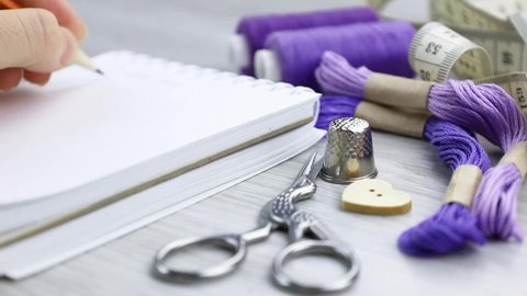 A woman writes in a notebook, a sewing plan.Purple spools of thread on the table.Sewing accessories.Sewing workshop.A white notebook for writing.