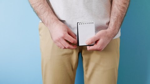 Diseases of the male reproductive system. A man in beige jeans holds a notebook with a question mark in two hands. Close up. Blue background. The concept of artificial insemination and impotence
