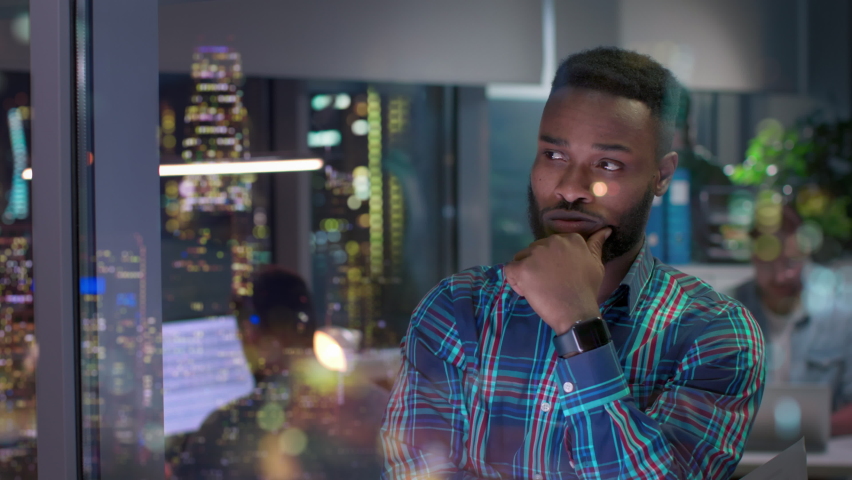 Stressed African-American businessman looking at night cityscape in office downtown. Pensive entrepreneur standing near window with illuminated skyscrapers reflection