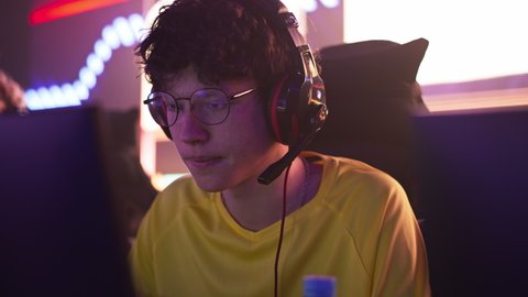 Focused teenager esportsman game player in glasses playing video game and speaking with teammates via headset during professional championship