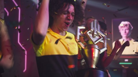 Professional esport team of gamers winners with curly hair men in front holding golden cup screaming and dancing amidst teammates while celebrating victory in gaming championship together