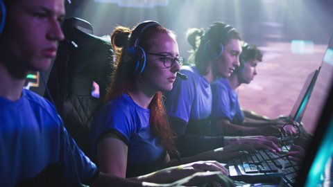 Concentrated young female esportsman gamer in glasses playing shooter on computer while sitting amidst teammates during gaming tournament