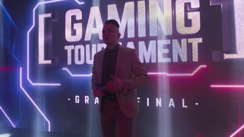 Zoom out tracking shot of adult male announcer walking under neon illumination and speaking at start of professional gaming final tournament championship