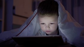 Small blond boy kid experiences emotion fear, covers his face with his hands while watching scary movie on laptop, lying at night under white blanket at night home on bed. Child is afraid scary movie.