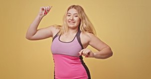 Happy overweight female dancing energetically. Cheerful blond plump woman in pink activewear smiling and looking at camera while dancing against yellow background
