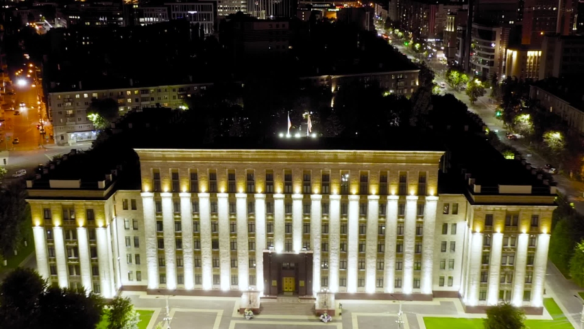 Voronezh, Russia. The House of the Soviets, a monumental Stalinist Empire style administrative building erected in 1953 during the post-war reconstruction of the city, dominates Lenin Square. The structure features a long symmetrical facade with tall vertical columns dramatically illuminated by warm golden and cool white uplighting against the night sky. The building sits at the center of a formal paved plaza with manicured green lawns, while the surrounding cityscape reveals a mix of mid-century and modern architecture along illuminated avenues with active traffic. The camera slowly pulls back, revealing the full scale of the landmark and its integration into the dense urban fabric. City night view, Aerial View, Departure of the camera. Dolly zoom.