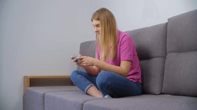 Gamer woman playing video game on smartphone. Excited white female in orchid polo shirt plays modern games on modern mobile phone device while sitting on couch in living room at home