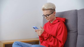 Tomboy woman using phone on couch in living room. Young white female with short dyed hair browsing internet on modern smartphone device. Person messaging online on social media app in 4k stock video