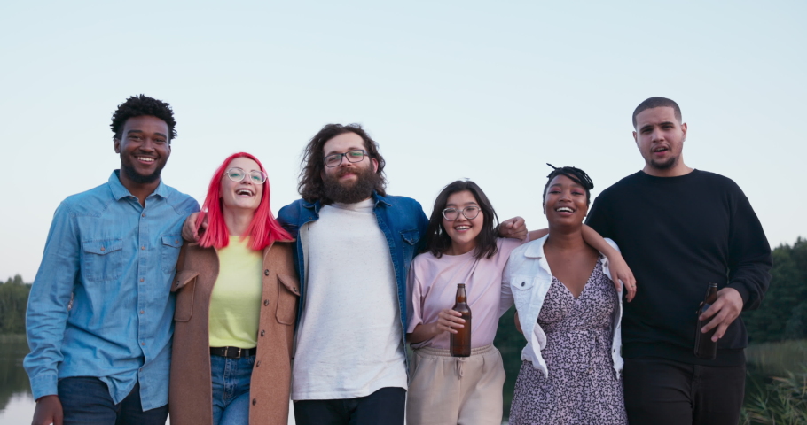 A group of best friends of different nationalities embrace in a row holding bottles in their hands, six smiling people lean forward at same time looking at camera, in the background a view of lake