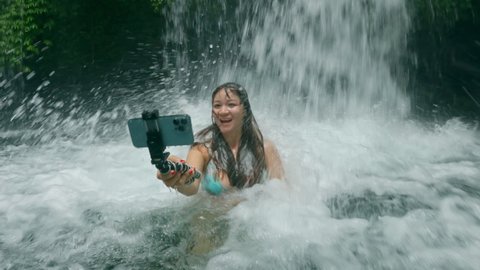 Crazy funny female blogger jumping in water under big waterfall in tropical river. Asian girl with white skin making selfie under water flow using a modern cell phone camera. Woman having fun in Bali
