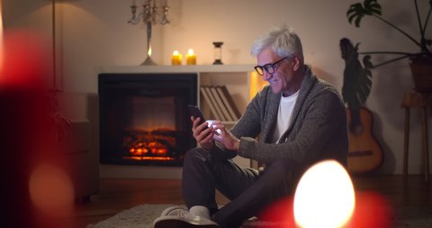 Happy senior man using smartphone while relaxing at home near fireplace. Portrait of aged male sitting on floor in living room and surfing internet on mobile phone enjoying evening at home