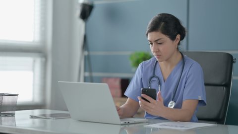 Indian Female Doctor using Smartphone while using Laptop in Office 