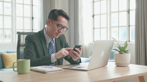 Asian Businessman With Glasses Wearing Business Suit Using Mobile Phone While Using Computer For Working At Home. 
