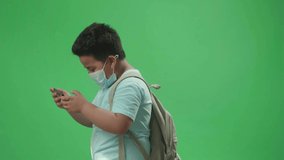 Side View Of Asian Boy Student Wearing A Mask And Playing Video Game On Mobile Phone While Walking To School On Green Screen Chroma Key

