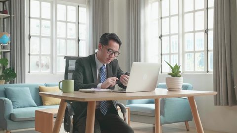 Asian Businessman Wearing Glasses And Business Suit Answering The Phone While Using The Computer For Working At Home.
