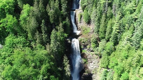 A Multiple Step Waterfall Is In View Between Tall Green Trees. This aerial view slowly backs away from the main subject throughout the video clip.
