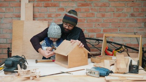 Dad and his little son are building a wooden bird feeder together in a home workshop. Father teaches boy how to hammer nails