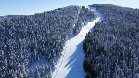 Aerial moving backward drone footage of skiers and snowboarders on Kopaonik mountain. Ski hill on a sunny day. Skiers and snowboarders skiing on snow slopes with ski lift.