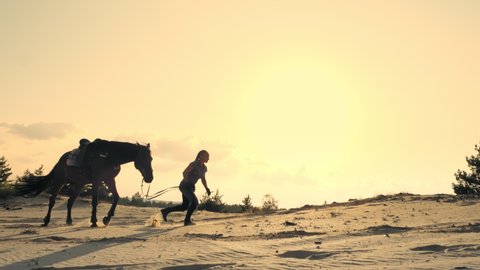 Horse riding. Silhouette of horsewoman, running with a horse, along the horizon of towering sandy hill, at sunset, on sky background and sun rays backlight.