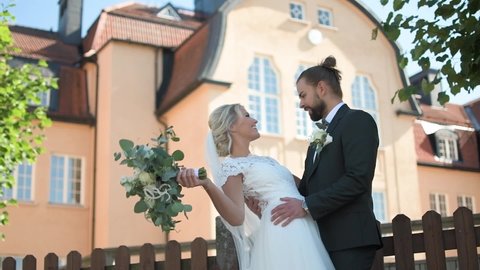 Happy wedding couple that are kissing behind wedding bouquet in front of a school building. Young caucasian bride and groom. Newlyweds. Bride in wedding dress. Bride Groom in jacket. Slow motion.