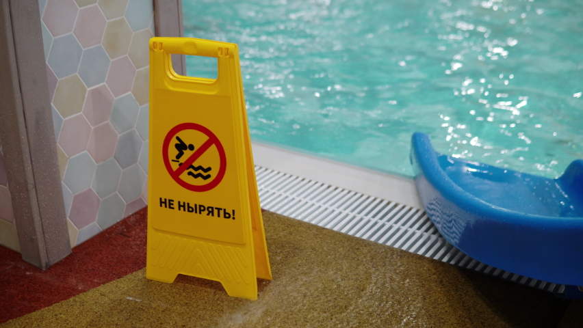 The inscription in Russian do not dive. A yellow sign stands near the pool with a warning inscription against the background of people floating in the pool 4k | Shutterstock HD Video #1086197201