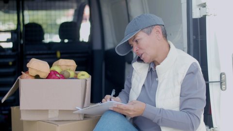 Courier talking with customer on phone in car, writing on tablet. Medium shot of woman in cap checking quantity of goods, preparing for delivering cardboard box of vegetables. Food delivery concept